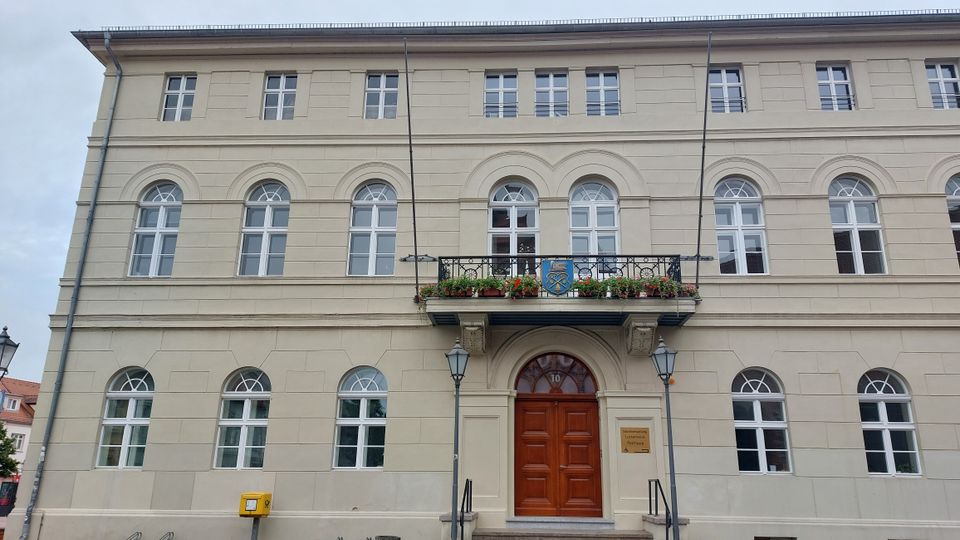 Historisches Gebäude mit hellbeiger Fassade, symmetrischen Rundbogenfenstern und einem hölzernen Eingangstor. Über dem Eingang befindet sich ein kleiner Balkon mit Blumen und einem Wappen, flankiert von zwei Laternen.