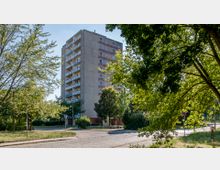Ein mehrstöckiges Wohngebäude aus Beton mit Balkonen auf jeder Etage, umgeben von Bäumen und Grünflächen. Im Vordergrund ist eine gepflasterte Straße sowie ein kleiner Platz mit Verkehrszeichen sichtbar.