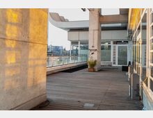 Eine Außenterrasse mit Holzboden, flankiert von einem modernen Gebäude mit Glasfassaden und Betonpfeilern. Im Hintergrund sind ein Geländer mit Glasbrüstung und eine urbane Skyline sichtbar.