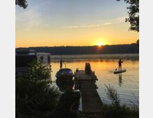 Ein ruhiger See bei Sonnenaufgang mit einem Holzsteg, einem geparkten Boot und einem Wohnmobil am Ufer; rechts paddelt eine Person auf einem Stand-up-Paddleboard über das Wasser, umgeben von Bäumen und vegetationsreicher Landschaft.