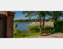Uferweg an einem See mit Schilfgürtel, Bäumen und einer Holzbank im Vordergrund; links ist ein Ziegelmauerstück zu sehen. Auf dem Wasser liegen mehrere Boote nahe einem Steg, im Hintergrund verläuft eine bewaldete Uferlinie unter blauem Himmel.