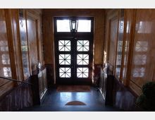 Treppenhaus mit einer dunkelbraunen Holztür im Eingangsbereich, die mit verglasten Fenstern in geometrischem Muster versehen ist. Die Wände sind mit glänzendem Marmor verkleidet, und dekorative Geländer aus Metall flankieren die Treppe.