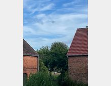 Backsteingebäude mit roten Ziegeldächern und einem grünen Baum in der Mitte des Bildes; im Hintergrund eine Wiese mit verschwommenen Personen, darüber ein blauer Himmel mit einigen weißen Wolken.