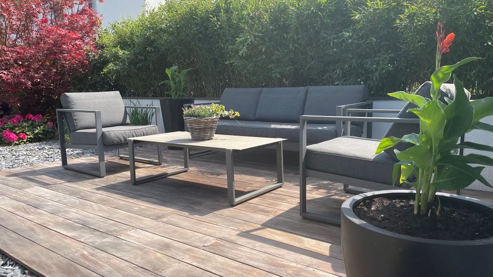 Holzterrasse im Garten mit modernen grauen Loungemöbeln und einem niedrigen Couchtisch. Im Hintergrund dichte grüne Hecke und ein roter Zierbaum, vorne ein großer Pflanzkübel mit hoher Pflanze und orangefarbener Blüte.