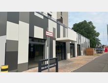 Moderne Gewerbefassade mit vertikalen Elementen in Grau- und Weißtönen. Vor dem Gebäude befinden sich eine gepflasterte Fläche, ein Straßenschild mit der Adresse „Allee der Kosmonauten 25A“ und ein Schild mit der Aufschrift „Fläche für die Feuerwehr“.