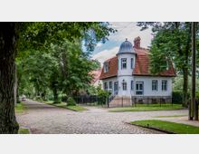 Weißes Einfamilienhaus mit roten Dachziegeln und einem runden Erker mit Kuppeldach, umgeben von einem Garten mit Metallzaun. Das Haus liegt an einer von Bäumen gesäumten, gepflasterten Straße in einer ruhigen Wohngegend.