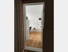 Blick durch eine geöffnete weiße Tür in einen hellen Raum mit Holzfußboden. Im Raum befinden sich ein kleiner Holztisch mit einem schwarzen Gerät darauf und ein schwarzer Stuhl.