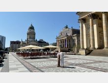 Weitläufiger Platz mit gepflastertem Boden, flankiert von historischen Gebäuden. Links die Kuppel des Deutschen Doms, rechts Säulen des Französischen Doms, in der Mitte ein Gebäude mit klassischer Architektur, Sitz des Konzerthauses Berlin, sowie Sitzbereiche mit Sonnenschirmen.