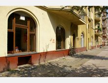 Gelbes Wohngebäude in einer städtischen Umgebung mit großen, bogenförmigen Fenstern im Erdgeschoss und roten Fassadenabschlüssen; im Vordergrund ein Fahrrad angelehnt an die Wand und ein mit Kopfsteinpflaster versehener Gehweg.