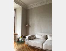 Wohnzimmer mit einer beigefarbenen Polstercouch auf Holzdielenboden, einem gläsernen Beistelltisch mit Blumen und einer Stehlampe mit konischem Schirm. Die Wand ist schlicht verputzt, oben mit dekorativer Stuckverzierung.