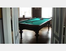 Ein Billardtisch mit grünem Filz steht in einem Raum mit Holzboden und großen Fenstern, durch die Tageslicht einfällt. Die Tür im Vordergrund ist geöffnet, und an der Wand hinter dem Tisch ist Tapete mit Ornamenten zu sehen.