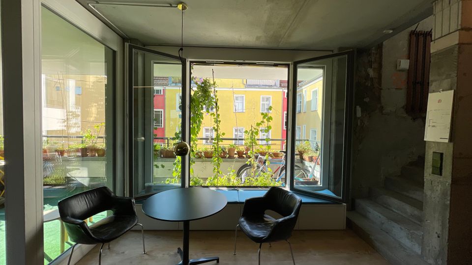 Ein moderner Innenraum mit Betonwänden, einer kleinen Wendeltreppe und einem großen Fenster, das auf einen Balkon mit Pflanzen und dahinterliegende bunte Wohnhäuser blickt. Zwei schwarze Stühle und ein runder Tisch befinden sich vor dem Fenster auf einem Holzboden.