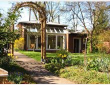 Ein Garten mit einem Holzbogen und Blumenbeeten führt zu einem kleinen Haus mit großen Fenstern und flachem Dach. Im Hintergrund sind Bäume und eine grüne Wiese sichtbar.