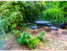 Ein gemütlicher Gartenbereich mit einem runden Metalltisch und vier passenden Stühlen, umgeben von üppigem Grün und bunten Blumenbeeten. Der Boden besteht aus einem Muster aus Steinpflaster und roten Ziegeln.