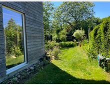 Ein Garten mit einer Rasenfläche, umgeben von blühenden Pflanzen und Sträuchern, im Hintergrund steht eine schwarze Gartenhütte unter einem großen Baum. Links im Bild ist ein modernes Holzhaus mit einer großen Fensterfront zu sehen.