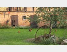 Ein Bauernhof mit einer roten Backsteinfassade, Holztüren und einem kleinen Fenster. Im Vordergrund sind ein Baum, ein altes Wagenrad und mehrere Hühner auf einer grünen Wiese zu sehen.