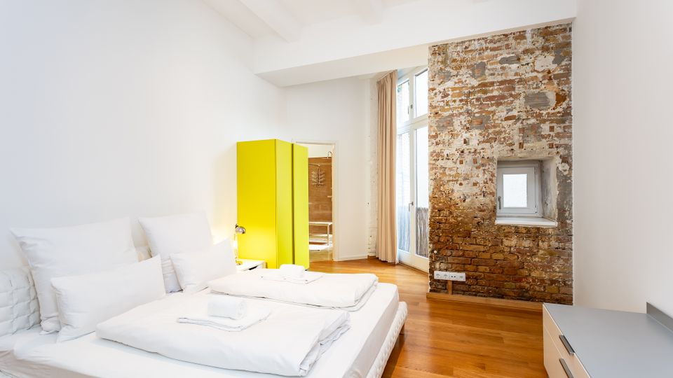 Helles Schlafzimmer mit Doppelbett, weißer Bettwäsche und Holzboden, kombiniert mit einer freiliegenden Backsteinwand mit kleinem Fenster. Ein auffälliger gelber Kleiderschrank steht neben bodentiefen Fenstern mit Vorhängen, die viel Tageslicht in den Raum lassen.