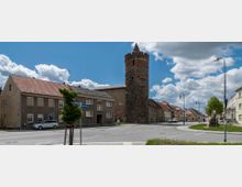 Straßenszene in einer Kleinstadt mit einem erhaltenen mittelalterlichen Rundturm aus dunklem Stein im Vordergrund, umgeben von Wohnhäusern mit roten Ziegeldächern. Eine Kreuzung mit Straßenschildern und Laternen ist gut sichtbar, während im Hintergrund ein bewölkter Himmel zu sehen ist.