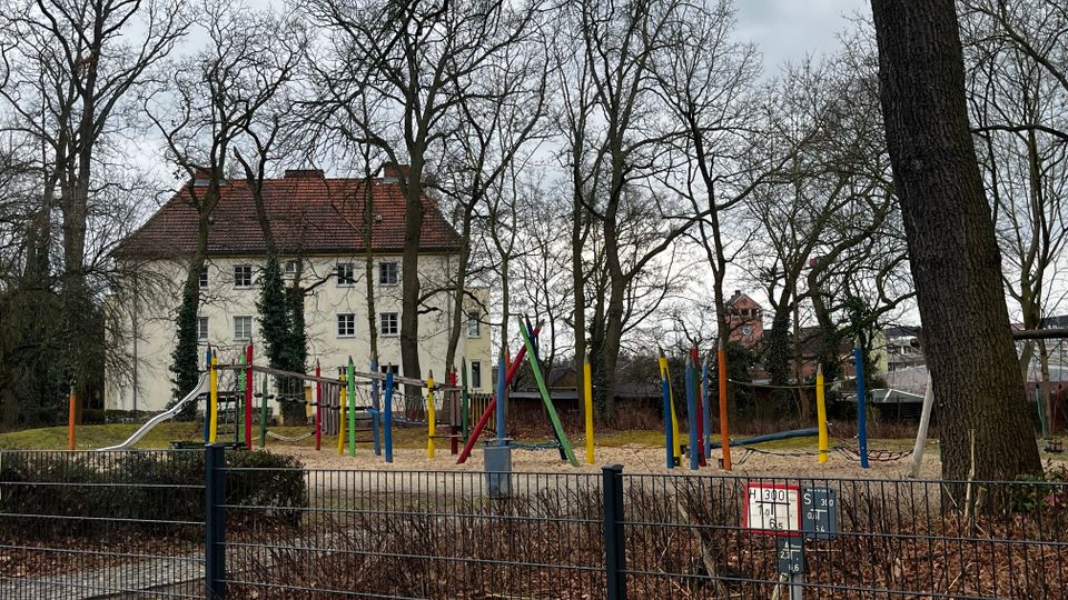 Eingezäunter Spielplatz mit Sandfläche, Rutsche und Klettergerüsten aus bunten Pfosten, umgeben von vielen kahlen Bäumen. Im Hintergrund steht ein helles Gebäude mit rotem Ziegeldach sowie weitere Häuser.