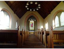 Innenansicht einer kleinen Kirche mit hölzernen Bänken, einem Ziegelfußboden und einem hohen, dunklen Holzgewölbe. Im Hintergrund befindet sich ein farbiges Glasfenster mit religiösen Motiven, umgeben von dekorativen Mustern an der Wand.