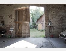 Innenansicht einer rustikalen Scheune mit Blick durch ein halb geöff­netes Holztor auf einen grünen Hof. Draußen sind Backsteingebäude sichtbar, drinnen befinden sich ein Waschbecken, ein Grill sowie elektrische Anschlüsse an der Ziegelwand.