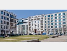 Ein moderner städtischer Platz mit Grünflächen, umgeben von mehrstöckigen Gebäuden aus heller Fassade mit großen Fenstern. Vor den Gebäuden sind parkende Autos, Fahrräder und einige Bäume sichtbar, während zwei Personen auf einer Parkbank sitzen.