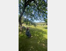 Eine Person schaukelt an einem Seil unter einem großen Baum auf einer Wiese. Im Hintergrund sind weitere Menschen auf Stühlen sowie eine weite, offene Landschaft mit Feldern und Bäumen zu sehen.