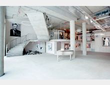 Ein moderner Galerieraum mit Betonwänden, freiliegenden Rohren an der Decke und mehreren großen Säulen. Die Galerie zeigt Fotografien und Kunstwerke an den Wänden, mit einer offenen Treppe, die zu einer Zwischengalerie führt. Im Vordergrund steht ein weißer Flügel mit Kerzen darauf.