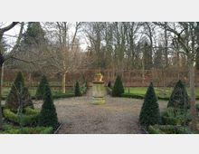 Ein formaler Garten mit symmetrischer Anordnung von Buchsbaumhecken und kegelförmigen Koniferen, die einen zentralen Steinsockel mit einem Pflanzentopf umgeben; im Hintergrund eine von kahlen Bäumen gesäumte Hecke.