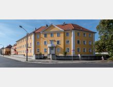 Gelbes Wohngebäude mit zwei Stockwerken und einem Satteldach, symmetrisch angeordneten Fenstern und einem kleinen Balkon über dem Hauseingang. Das Gebäude liegt an einer Straßenkreuzung, umgeben von Gehwegen, Bäumen und weiteren Gebäuden.