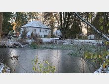 Ein gefrorener Teich in einer winterlichen Landschaft mit leichtem Schneefall, umgeben von Bäumen und Sträuchern. Im Hintergrund ist ein weißes zweistöckiges Gebäude mit Satteldach und ein weiterer Gebäudeausschnitt sichtbar.