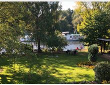 Grüne Parklandschaft mit Rasen, Blumenbeeten und Bäumen, im Hintergrund ein Fluss mit einem weißen Ausflugsboot. Rechts befindet sich eine überdachte Sitzgelegenheit, und vorne stehen Gartenmöbel auf der Wiese.