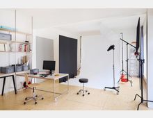Ein modernes Fotostudio mit hellem Holzfußboden, ausgestattet mit einer weißen Hohlkehle, Beleuchtungsstativen und Reflektoren. Im Vordergrund steht ein Schreibtisch mit einem Monitor, einem drehbaren Stuhl und Regalen mit Zubehör an der Wand.
