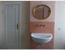 Weiße Wand mit einer kleinen Tür auf der linken Seite und einer beige-rosafarbenen Fläche rechts, an der ein wandmontiertes Waschbecken, ein goldgerahmter ovaler Spiegel und silberne Armaturen zu sehen sind.