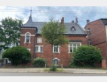 Backstein-Gebäude mit steilem Schieferdach, Ornamenten an der Fassade und großen Fenstern, umgeben von Bäumen und Sträuchern, mit einem Metallzaun zur Straße hin. Ein kleiner Baum steht im Vordergrund neben dem Gehweg.