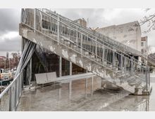 Betongebäude mit einer breiten Außentreppe und Metallgeländern, die zu einer oberen Ebene führt. Im Erdgeschoss befinden sich große Fenster mit Spiegelflächen, daneben stehen weiße Gartenmöbel und Pflanzentöpfe; die Umgebung zeigt eine urbane Straße mit kahlen Bäumen.