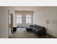 Schlichtes Wohnzimmer mit weiß gestrichenen Wänden, einem grauen Ecksofa, einer Stehlampe und zwei großen Fenstern, die Tageslicht hereinlassen. Ein schmaler Holztisch steht an der Wand, daneben führt eine offene Tür in einen angrenzenden Raum.