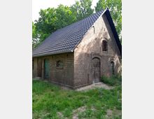 Backsteingebäude mit Satteldach aus dunklen Ziegeln, umgeben von Gras und Bäumen. Das Gebäude hat mehrere kleine Fenster, eine grüne Holztür und eine größere Holztoröffnung auf der rechten Seite.