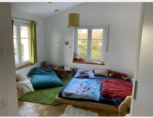 Ein gemütliches Kinderzimmer mit einem niedrigen Bett, bunten Bettdecken und einem großen Sitzsack auf einem grünen Teppich. Die Wände sind weiß, das Zimmer hat zwei große Fenster mit grünen Vorhängen, ein Holzregal mit Büchern und Spielzeug, sowie Ausblick auf einen Garten.