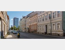 Straßenszene mit Altbauten auf der rechten Seite, teils mit bröckelnden Fassaden, und einem modernen Hochhaus im Hintergrund. Auf dem Gehweg stehen ein gelbes Motorrad und geparkte Autos, während eine Straßenbahnlinie die Straße überquert.