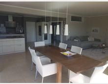 Offener Wohnraum mit modernem Design, bestehend aus einer Küche, einem Essbereich und einer Sitzecke. Im Vordergrund steht ein Holztisch mit weißen Stühlen und hängenden Lampen darüber, im Hintergrund befinden sich eine graue Couch, ein Teppich und Küchenschränke.