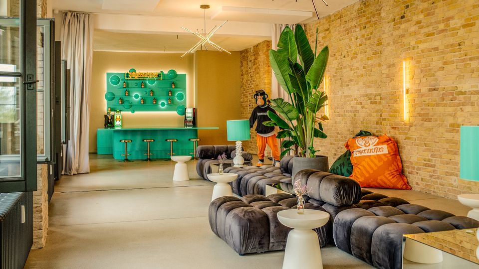 Moderner Lounge- und Barbereich in einem Loft mit Sichtziegelwand, großen schwarzen Polstersofas und kleinen weißen Beistelltischen. Im Hintergrund steht eine türkisfarbene Bar mit Hockern und beleuchtetem Flaschenregal; dekorative Deckenleuchten, hohe Pflanzen und Vorhänge rahmen den Raum.