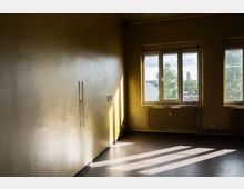 Leerer Innenraum mit beigefarbenen Wänden und dunklem Boden, von zwei großen Fenstern durch Tageslicht stark ausgeleuchtet. Unter den Fenstern befinden sich Heizkörper, und durch die Scheiben sind Bäume und Gebäude im Außenbereich zu sehen.