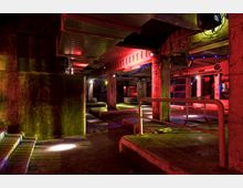 Industrieähnlicher Innenraum mit rohen Betonpfeilern, Deckenstreben und metallenen Geländern, in rot und gelb-grün beleuchtet. Der Raum wirkt schummrig und verfügt über bunt beleuchtete Bereiche, die eine Club- oder Veranstaltungsatmosphäre andeuten.