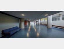 Weitläufiger, moderner Flur mit blauem Bodenbelag und runden Betonsäulen. Links eine blaue Sitzbank an der Wand, rechts Geländer und große Fenster, die Tageslicht einfallen lassen.