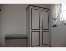 Ein kleiner Raum mit einem schlichten weißen Kleiderschrank mit blauen Verzierungen und zwei Türen sowie einer Fensterfront mit weißen, blau verzierten Vorhängen. Links neben dem Schrank befindet sich eine kleine Ablagefläche.
