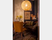 Gemütliche Sitzecke in einem Café oder Wohnraum mit dunkler Eckbank und Kissen, Holztisch und senfgelben Polsterstühlen auf gemustertem Fliesenboden. An der dunkelblauen Wand hängen gerahmte Bilder, darüber eine große runde Pendelleuchte; auf dem Tisch steht eine Vase mit gelben Blumen.