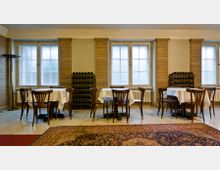 Ein heller Raum mit mehreren kleinen Tischen, die von Holzstühlen umgeben sind, und großen Fenstern an der Rückwand. Zwischen den Fenstern stehen schmale Regale mit aufbewahrten Weinflaschen, und der Boden ist mit Teppichen dekoriert.