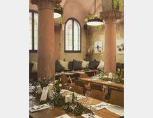 Ein stilvoll dekorierter Innenraum eines Restaurants mit hohen, verzierten Säulen und gewölbter Decke. Die Tische sind mit Gläsern, Menükarten, weißen Blumen und grünen Pflanzenarrangements geschmückt, während durch große Rundbogenfenster diffuses Licht einfällt.
