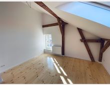 Dachgeschossraum mit schräger Decke, sichtbaren Holzbalken, einem großen Dachfenster und hellem Holzboden; an der weißen Wand befindet sich ein schmales Fenster mit Heizkörper darunter.